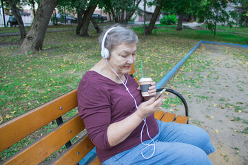 Elderly woman with headphones and cup of coffee sits on bench and talks on the phone with her...