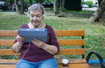 Elderly woman with headphones sits on bench and talks on a tablet. The concept of  adaptation of...