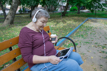 Elderly woman with headphones and cup of coffee sits on bench and talks on a tablet. The concept of...