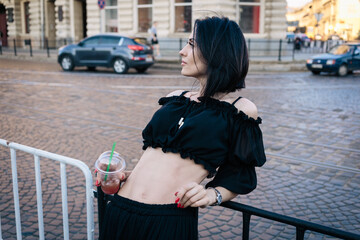 A beautiful girl with short black hair in a black top and skirt stands near the road, leaning against a fence, holding a plastic glass with lemonade in her hand and looking at the road. downtown.