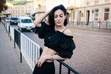 A beautiful girl with short black hair in a black top and skirt stands near the road, leaning against a fence, her hand on the hair and looking down. downtown. City. Cars on the background.