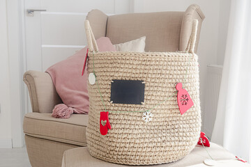 A jute handmade basket with a small black chalkboard and a red and white decorative wood garland on a pouf next to a beige armchair on a white wallpaper background. New year decoration concept