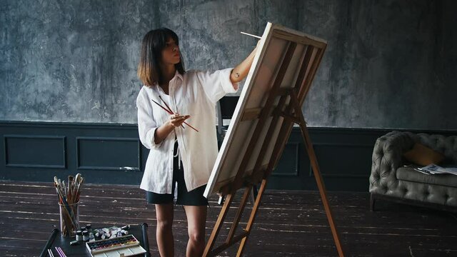 Young woman painter is drawing a picture on canvas using oil paints, palette and paintbrushes while working at her studio