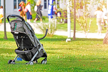 empty baby stroller on grass in city park landscape with crowd walking along street on background. Picnic in park. Autumn in town. Summer outdoor activity. Authentic lifestyle