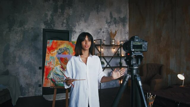 Woman painter posing near a portrait in modern art style against camera fixed on tripod and recording video for her followers