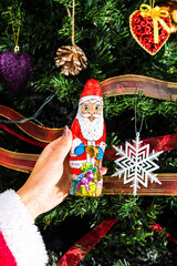 Hand holding Santa Claus chocolate figurine in front of the Christmas tree. Decorating Christmass...