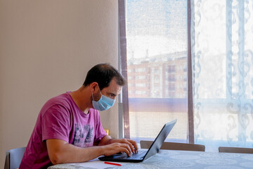 Fototapeta premium Worried bearded man dressed in a purple t-shirt and wearing a mask to protect himself from the coronavirus looking at the laptop