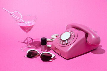 Old style rotary telephone on pink background with cocktail, sunglasses and nail polish. Minimal retro 80's concept. Flat lay.