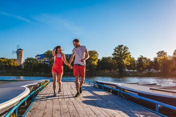 Young man and woman running along summer river dock. Couple having fun at sunset holding hands....