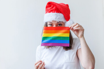 Beautiful lesbian girl in red Santa Claus hat with LGBT rainbow flag isolated on white background looking happy and excited. Young woman Gay Pride portrait. Happy Christmas and New Year holidays