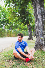 Young man dressed in sportswear performs stretching exercises sitting on the ground in a park. Lifestyle.