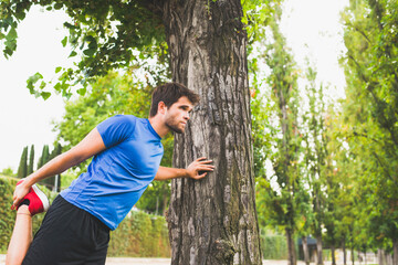Naklejka na ściany i meble Young man dressed in sportswear performs stretching exercises leaning on a tree in the park. Lifestyle.