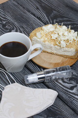 Cheesecake with white chocolate and a cup of coffee. The filling is visible on the cut of the dessert. Nearby is a medical mask and antiseptic. On pine planks painted black and white.