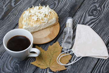 Cheesecake with white chocolate and a cup of coffee. The filling is visible on the cut of the dessert. Nearby is a medical mask, antiseptic and dried maple leaves. On pine planks