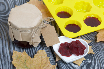 A jar of cherry jam with a label for an inscription and a saucer of cherry marmalade. A silicone mold with marmalade is visible. Dried maple leaves all around. On pine planks painted black and white.