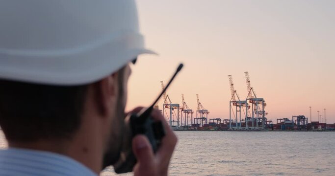 The man puts a white protective helmet on his head and speaks on the radio against the background of the seaport.