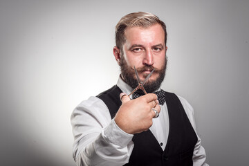 Portrait of stylish professional hairdresser with beard. Man wearing shirt, looking at camera and holding scissors near his beard