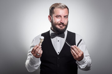 A handsome man in a shirt with a bow tie and a vest holding a blade, a brush with shaving foam. Professional barber concept