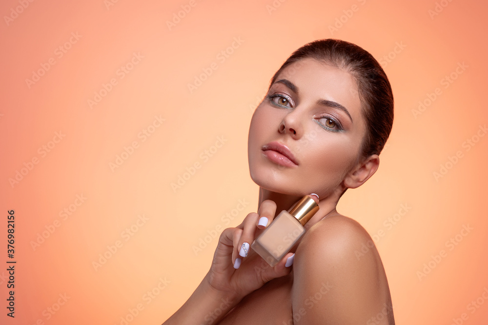 Wall mural Young brunette woman with naked shoulders, natural makeup and perfect clean fresh skin holding foundation bottle in hand. Studio portrait on orange background. Facial skincare and anti-age treatment