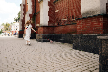 A young woman walks with a Scottish straight cat on the street on a leash in the city. Blonde girl dressed in a long white dress