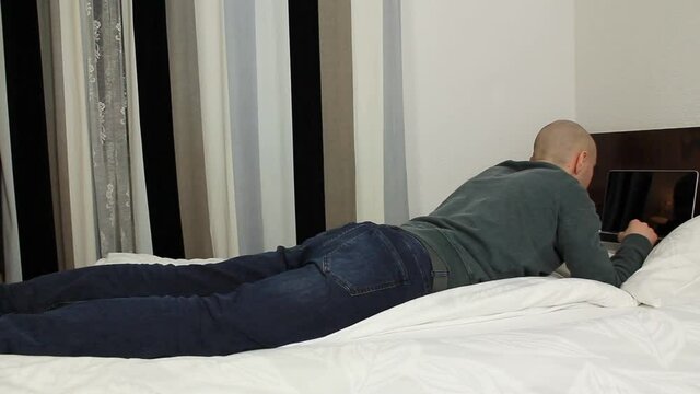 young bald man in a gray sweater and jeans lies on a bed in a hotel room, includes a laptop, the concept of business travel, tourism, loneliness, stay at home, quarantine