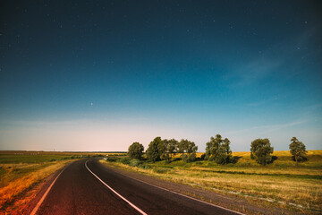 Blue Night Starry Sky Above Country Asphalt Road In Countryside And Green Meadow. Night View Of Natural Glowing Stars