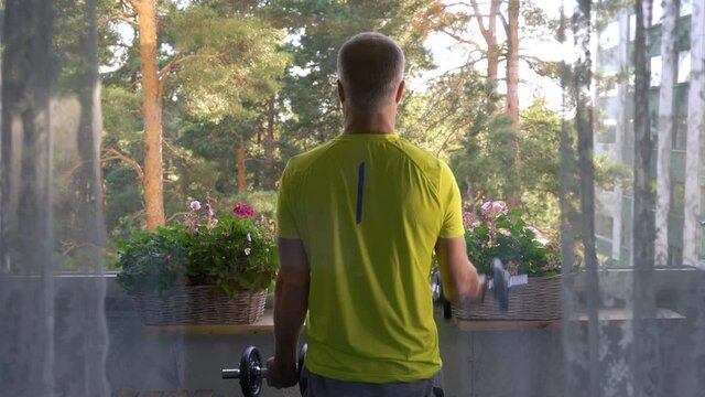 A man in sportswear playing sports with dumbbells on the balcony of his apartment during quarantine and self-isolation