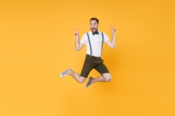 Fototapeta na wymiar Full length portrait of excited young bearded man 20s wearing white shirt suspender shorts posing jumping showing victory sign looking camera isolated on bright yellow color wall background studio.