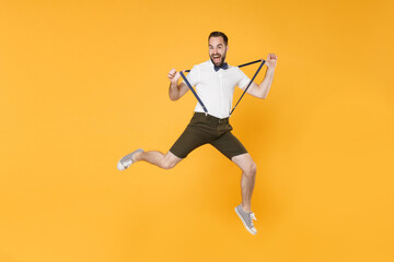 Fototapeta na wymiar Full length portrait of crazy screaming young bearded man 20s wearing white shirt shorts posing jumping stretching suspender looking camera isolated on bright yellow color wall background studio.