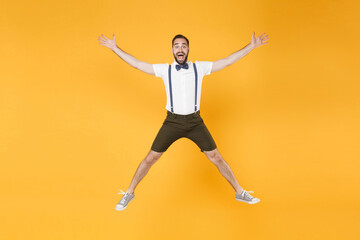 Fototapeta na wymiar Full length portrait of excited cheerful young bearded man 20s wearing white shirt suspender shorts posing jumping spreading hands and legs looking camera isolated on bright yellow background studio.