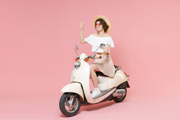 Fototapeta na wymiar Full length portrait of smiling young woman in white summer clothes hat glasses waving and greeting with hand as notices someone sitting driving moped isolated on pastel pink colour background studio.
