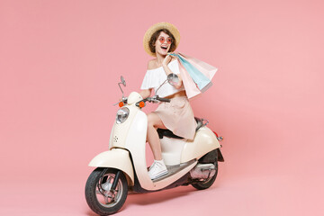 Obraz na płótnie Canvas Full length portrait of cheerful young woman in white clothes hat glasses hold package bag with purchases after shopping looking aside driving moped isolated on pastel pink colour background studio.