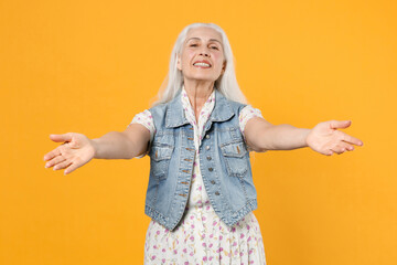 Smiling cheerful funny elderly gray-haired female woman 60s 70s wearing casual dress denim waistcoat posing reach out stretch hands looking camera isolated on yellow color background studio portrait.