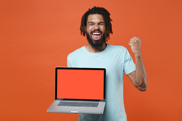 Happy young african american man guy in blue t-shirt isolated on orange background. People lifestyle concept. Mock up copy space. Hold laptop pc computer with blank empty screen doing winner gesture.
