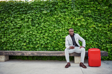 Businessman sitting on a bench with a red suitcase and mobile phone in hand.