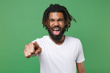 Fototapeta premium Angry young african american man guy with dreadlocks 20s wearing white casual t-shirt posing pointing index finger on camera screaming swearing isolated on green color wall background studio portrait.