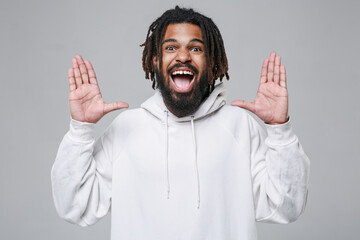 Excited surprised young african american man guy with dreadlocks 20s in white casual streetwear hoodie posing keeping mouth open spreading hands isolated on grey color wall background studio portrait.