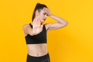Fototapeta na wymiar Dissatisfied young fitness sporty woman in black sportswear posing training working out showing stop gesture with palm put hand on head keep eyes closed isolated on yellow background studio portrait.