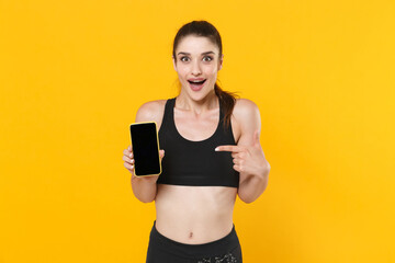Excited young fitness sporty woman in black sportswear posing training pointing index finger on mobile phone with blank empty screen mock up copy space isolated on yellow background studio portrait.