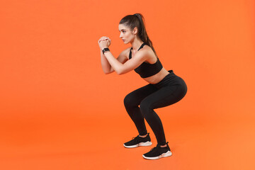 Full length side view portrait strong young fitness sporty woman in black sportswear training working out doing exercise squatting hold hands folded looking aside isolated on orange background studio.