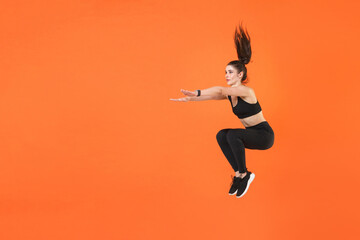 Fototapeta na wymiar Full length portrait of young fitness sporty woman 20s wearing black sportswear training jumping doing exercise squatting stretching hands looking aside isolated on orange color background studio.