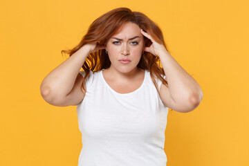 Obraz na płótnie Canvas Displeased sick tired young redhead plus size body positive female woman girl 20s in white casual t-shirt posing put hands on head having headache isolated on yellow color background studio portrait.