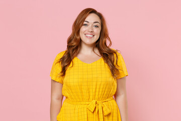 Smiling beautiful attractive young redhead plus size body positive female woman girl 20s in yellow casual dress posing looking camera isolated on pastel pink color wall background studio portrait.