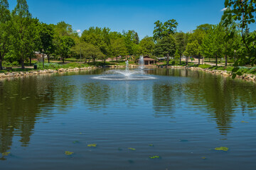 Fototapeta na wymiar Fountain aerators spray water on blue pond with a stone picnic stand in the background. Sun light reflects off of water droplets suspended mid air in this scenic setting.