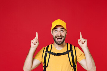 Delivery man 20s in yellow cap t shirt uniform thermal bag backpack points finger up head isolated...