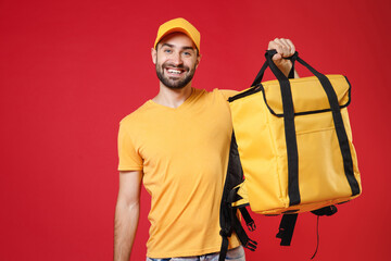 Delivery employee man guy 20s in yellow cap t-shirt uniform hold thermal bag backpack with food work as courier isolated on red background studio. Service during quarantine coronavirus covid-19 virus.