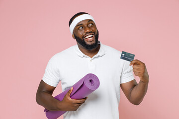 Pensive young bearded african american fitness sports man in headband t-shirt hold yoga mat credit bank card looking up spending time in gym isolated on pastel pink color background studio portrait.