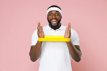 Excited young bearded african american fitness sports man 20s in white headband t-shirt doing exercise with fitness gums spending time in gym isolated on pastel pink color background studio portrait.
