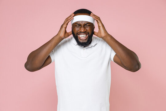Sick Displeased Young Bearded African American Fitness Sports Man In Headband T-shirt Having Headache Put Hands On Head Spending Time In Gym Isolated On Pink Color Wall Background Studio Portrait.