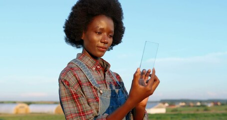 African American woman in field holding glass transparent device and tapping or scrolling on it. Interface of future. Futuristic technologies. Girl using gadget of hi-tech. Countryside concept.